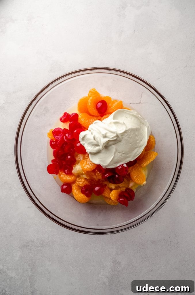 Aerial photo of canned pineapple, mandarin oranges, maraschino cherries, and sour cream in a bowl to make ambrosia salad.