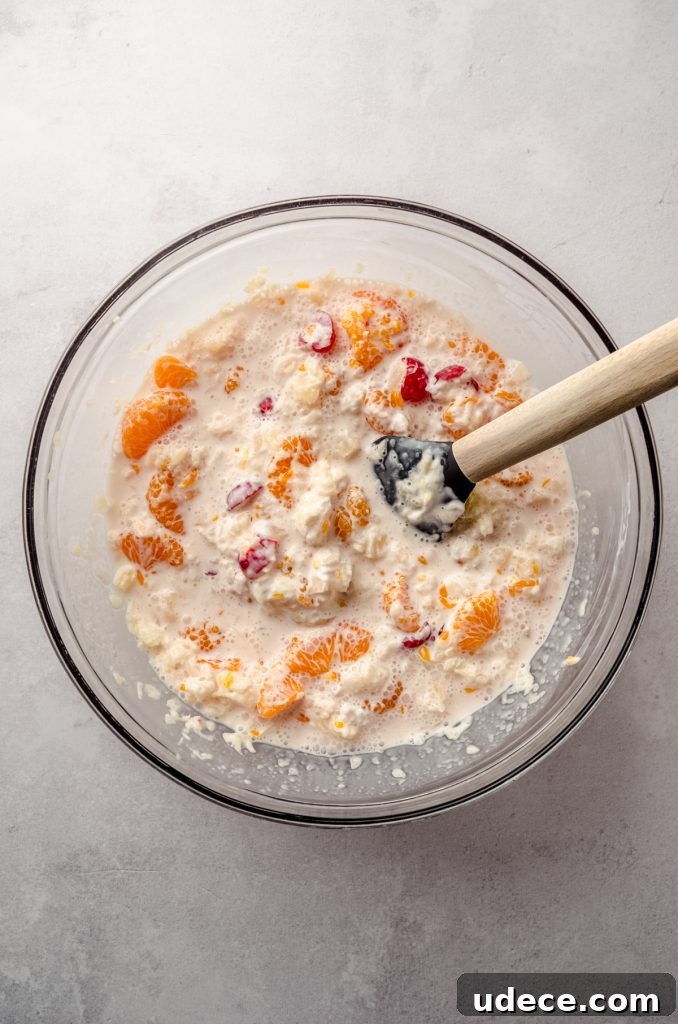 Aerial photo of canned pineapple, mandarin oranges, maraschino cherries, and sour cream in a bowl to make ambrosia salad.