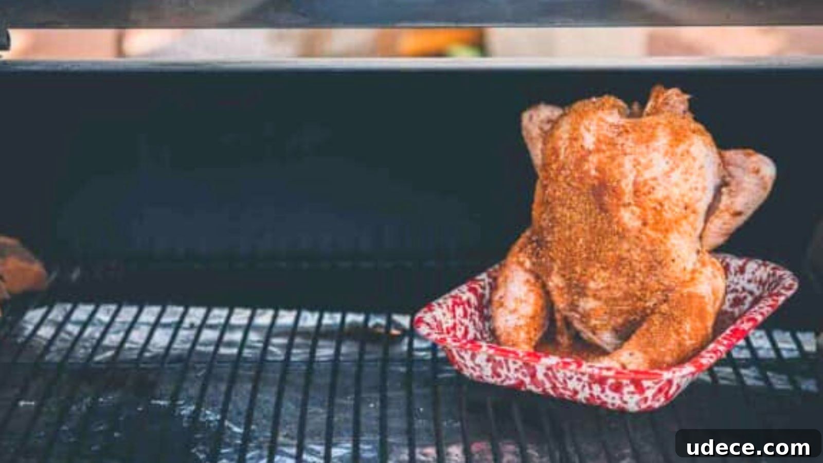 Whole chicken seasoned and placed upright in a red speckled roasting pan on a grill.