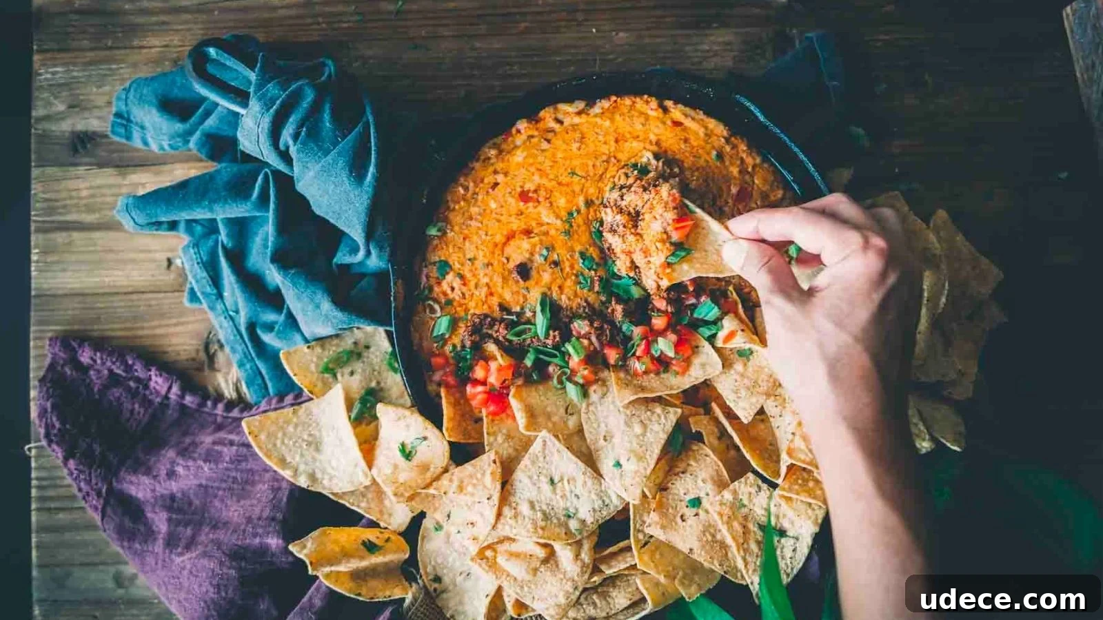 A hand dips a tortilla chip into a pan of cheesy, seasoned dip, surrounded by more chips on a wooden surface.