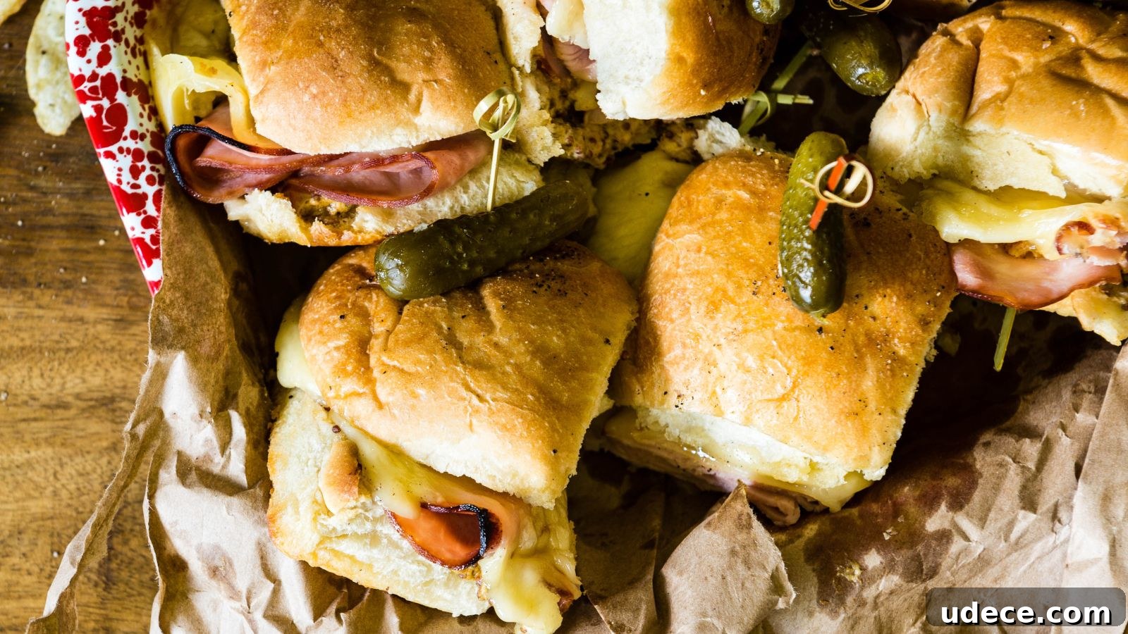 Close-up of ham and cheese sliders on crumpled parchment paper, each topped with a pickle and secured with a toothpick.