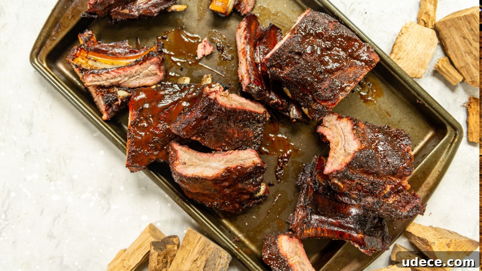 Barbecued beef ribs on a metal baking tray, surrounded by wood chips, against a light background.