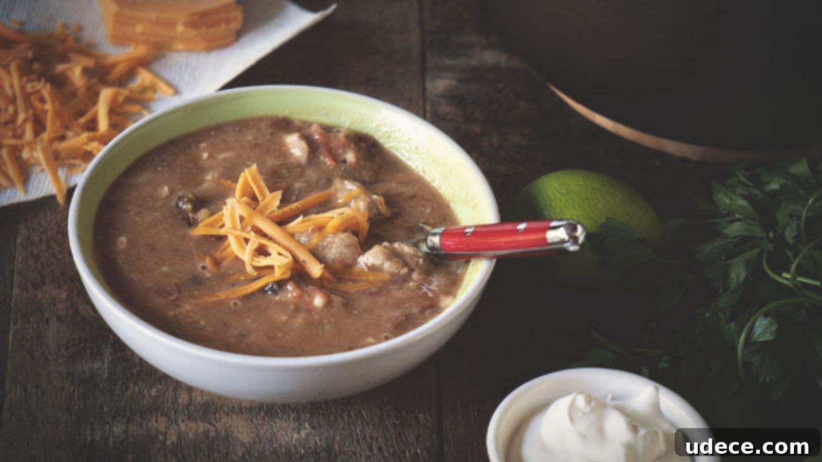 Bowl of soup with chunks of meat and vegetables, topped with shredded cheese. A red-handled spoon rests in the bowl. Surrounding items include shredded cheese, lime, herbs, and sour cream.