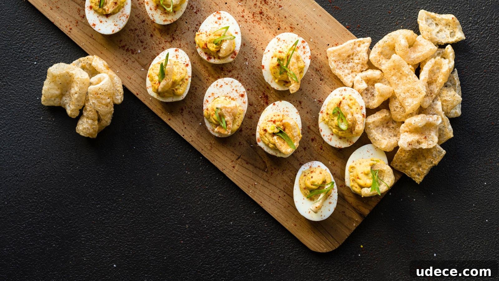 Deviled eggs on a wooden board garnished with spices and green herbs, accompanied by crispy pork rinds on a dark surface.
