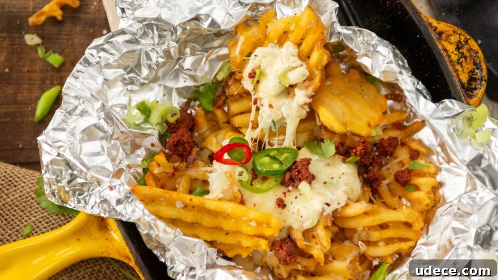 Waffle fries with melted cheese, red and green chili slices, and herbs, served in foil on a skillet.