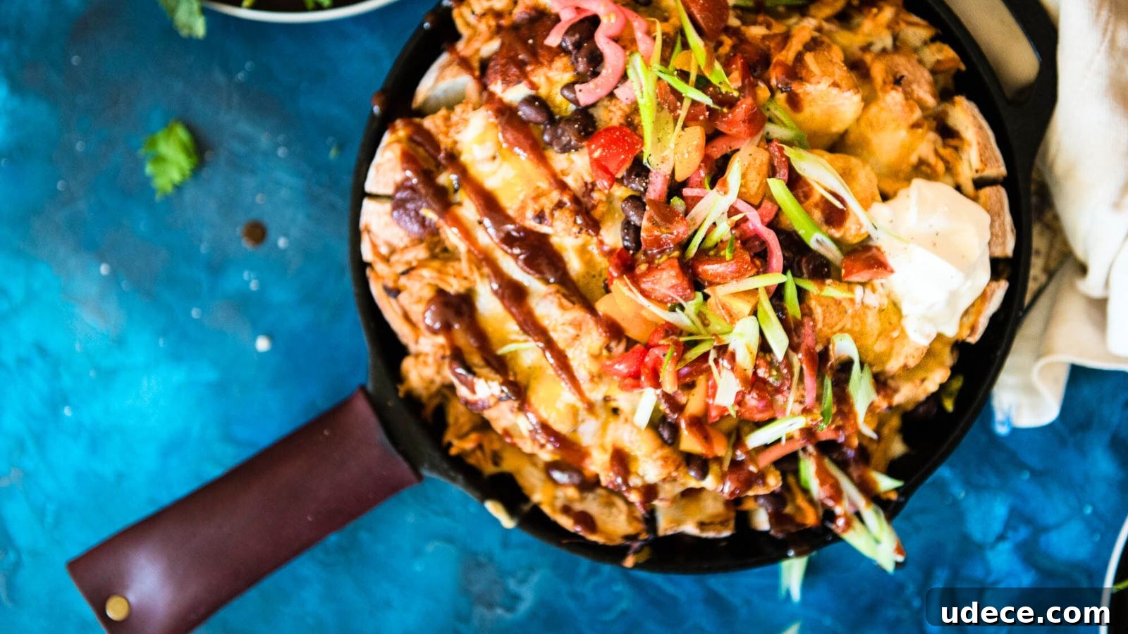 A skillet filled with loaded nachos topped with cheese, sour cream, sliced green onions, and diced tomatoes on a blue background.
