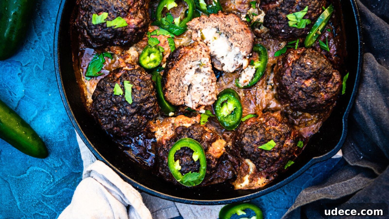 Meatballs in a pan with sliced jalapeños and garnished with herbs on a blue surface. Some meatballs are broken open.