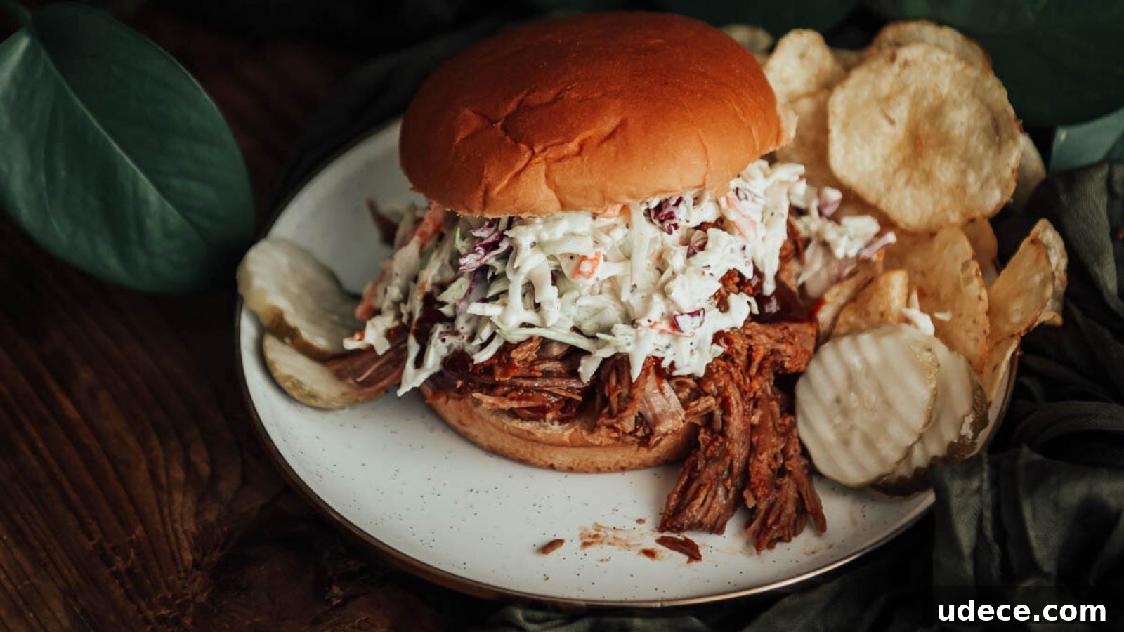Pulled pork sandwich with coleslaw on a bun, served on a white plate with potato chips and a pickle slice, set against a wooden table with green leaves in the background.