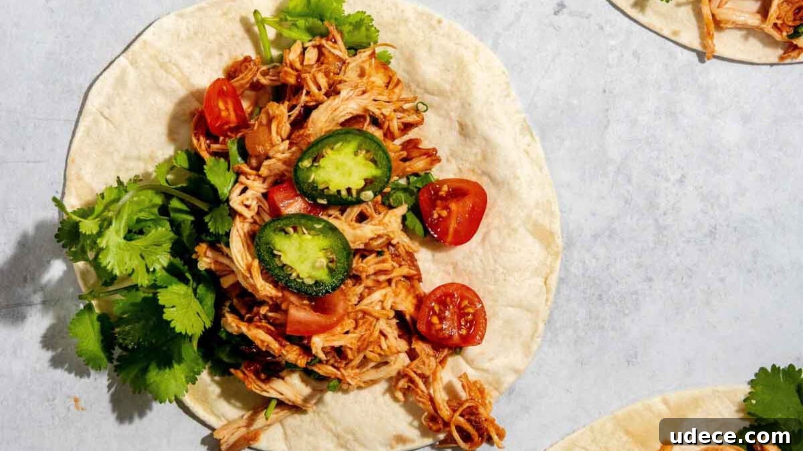 A tortilla with shredded chicken, sliced jalapeños, cherry tomatoes, and cilantro on top, placed on a light surface.