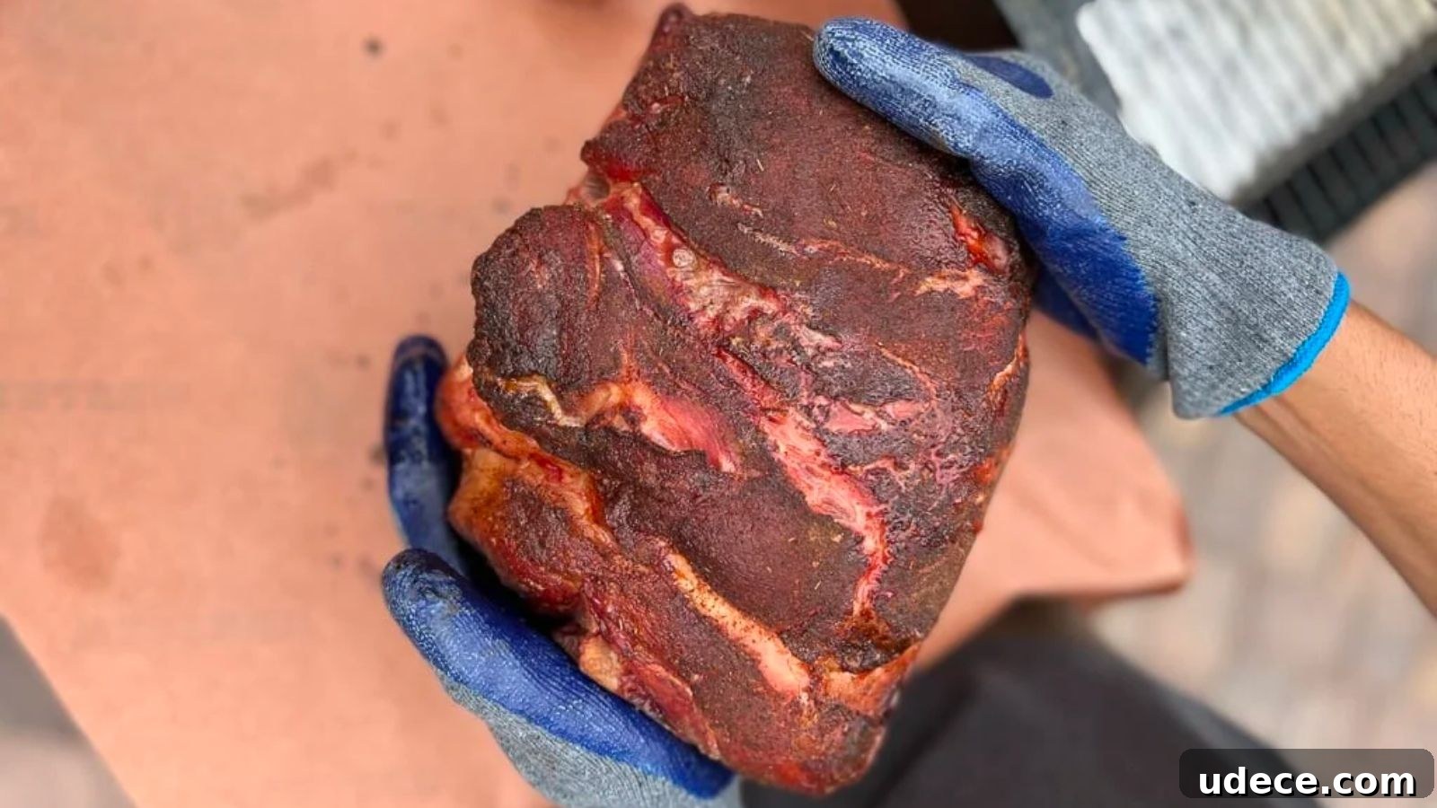 Gloved hands holding a large piece of seasoned, cooked meat on a brown surface.